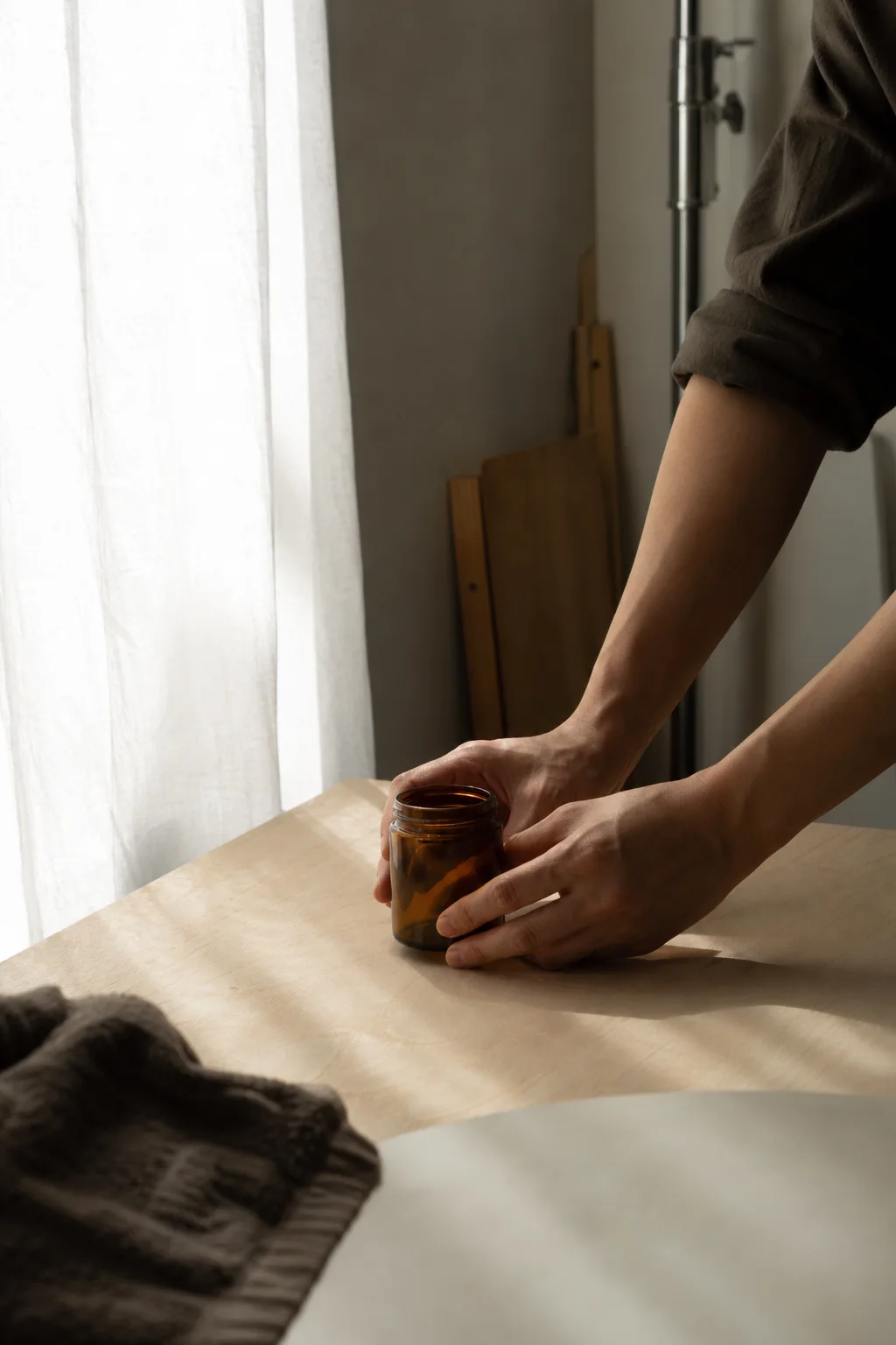 AI-generated editorial photograph of a maker placing an unlabelled amber jar beside a softly curtained window